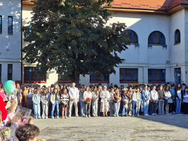 Un grup mare de persoane stă în fața unei clădiri albe cu ferestre și sub un copac mare.