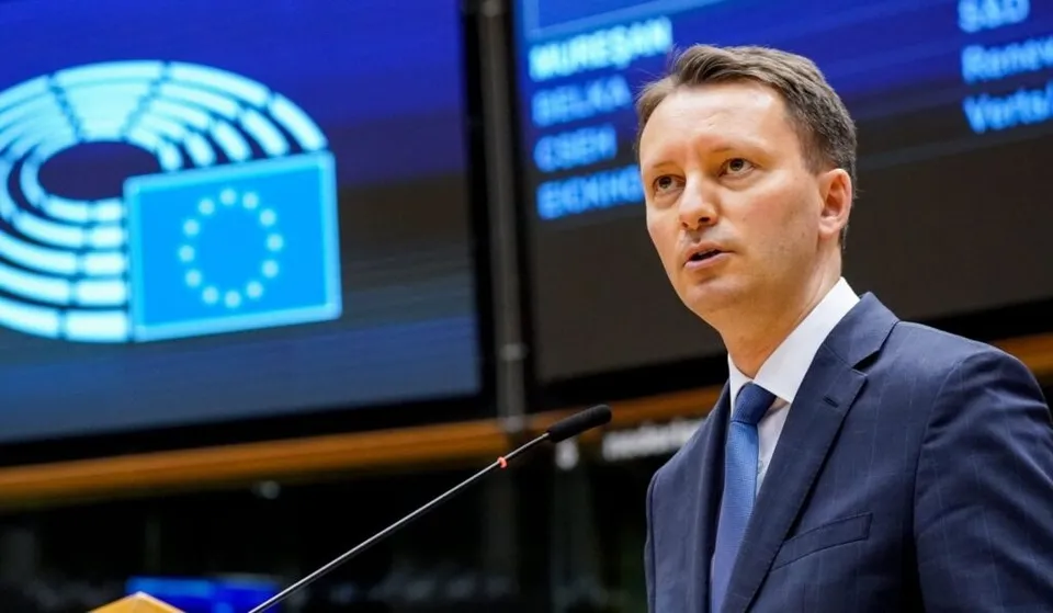 Un bărbat cu părul scurt, purtând sacou închis la culoare, cămașă albă și cravată albastră, stă în fața unui microfon într-un spațiu cu ecrane afișând simboluri.