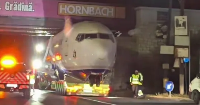 O componentă mare de avion este transportată pe un vehicul special sub un pod, lângă un panou publicitar de culoare roz.
