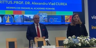 Un bărbat cu ochelari și cravată roșie stă în spatele unui podium, lângă o femeie cu părul lung și haine negre, în fața unui ecran albastru.