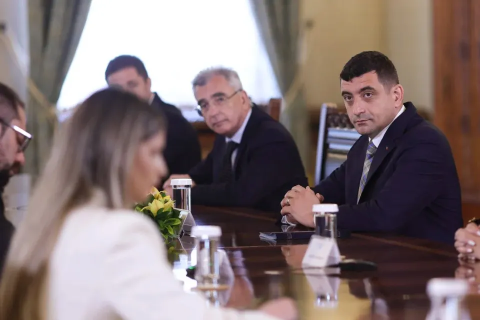 Patru persoane sunt așezate la o masă de conferințe într-o încăpere cu draperii, purtând haine formale și având pahare de sticlă în față.