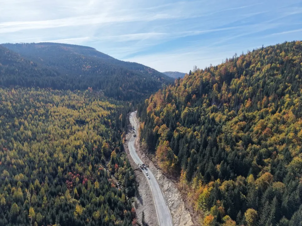 O drum de țară străbate o vale împădurită cu copaci în nuanțe de verde și galben, fiind înconjurat de versanți montani copaci.