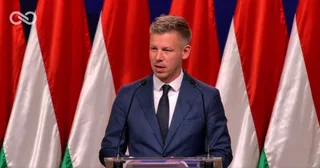 Un bărbat cu părul scurt și deschis la culoare poartă costum albastru, cămașă albă și cravată neagră în fața unor steaguri roșu și albe.