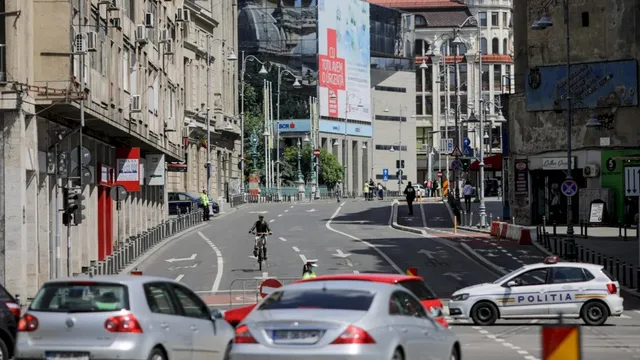 Mașini circulă pe un drum urban lângă clădiri înalte, în timp ce un biciclist pedalează pe banda de circulație și o mașină de poliție este parcată.