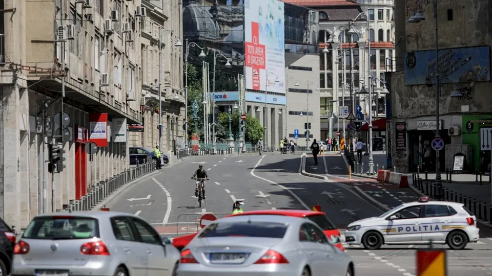 Mașini circulă pe un drum urban lângă clădiri înalte, în timp ce un biciclist pedalează pe banda de circulație și o mașină de poliție este parcată.