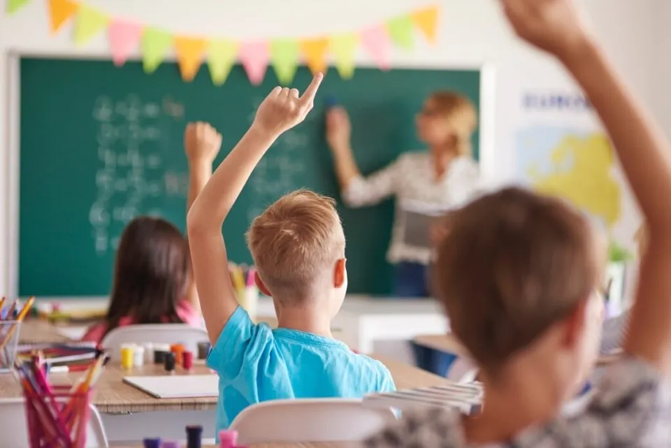 O sală de clasă cu copii ridicați la tablă, ghiozdane și materiale colorate, în fața unui profesor neclar.