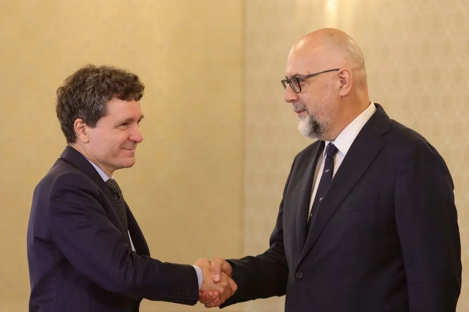 Un bărbat cu părul creț și un bărbat chel cu ochelari, ambii purtând costume în armonie, se strâng de mână în fața unui fundal neutru.