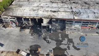 O clădire industrială cu acoperiș metalic deteriorat și porți de garaj distruse prezintă urme de ardere și resturi pe asfaltul din fața sa.