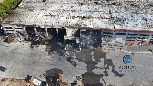 O clădire industrială cu acoperiș metalic deteriorat și porți de garaj distruse prezintă urme de ardere și resturi pe asfaltul din fața sa.