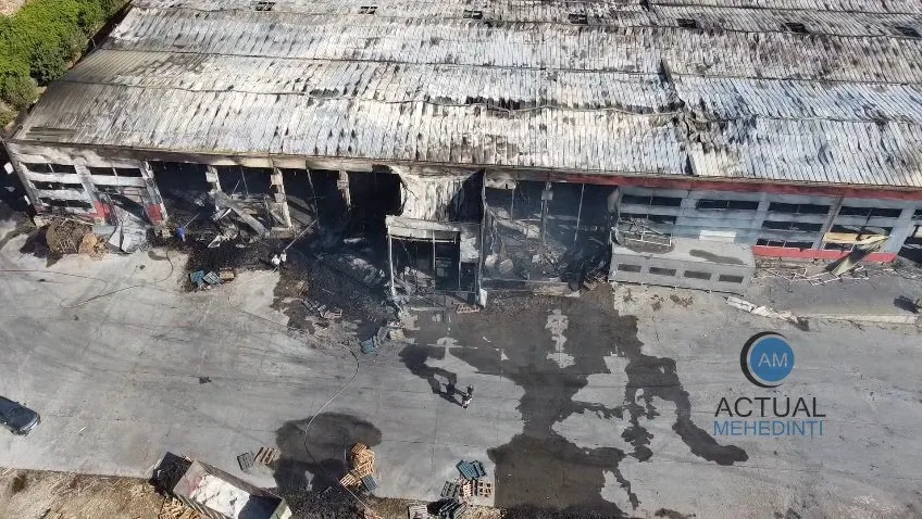 O clădire industrială cu acoperiș metalic deteriorat și porți de garaj distruse prezintă urme de ardere și resturi pe asfaltul din fața sa.