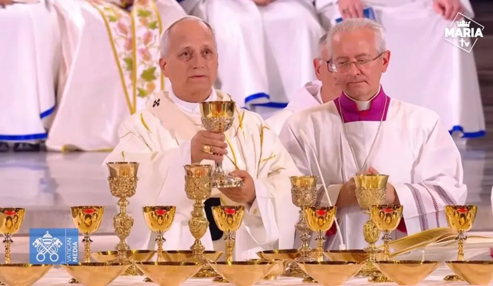 Un bărbat în robă albă ține un chalice aurit în fața mai multor chalice similare așezate pe o masă.