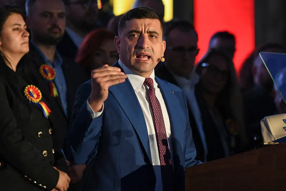 Un bărbat într-un costum albastru și cravată roșie vorbește în fața unui grup de persoane din spatele unui podium.