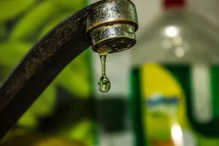O picătură de apă suspendată sub un robinet metalic într-un cadru cu fundal verde și un recipient de detergent neclar.