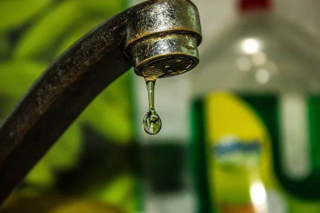 O picătură de apă suspendată sub un robinet metalic într-un cadru cu fundal verde și un recipient de detergent neclar.