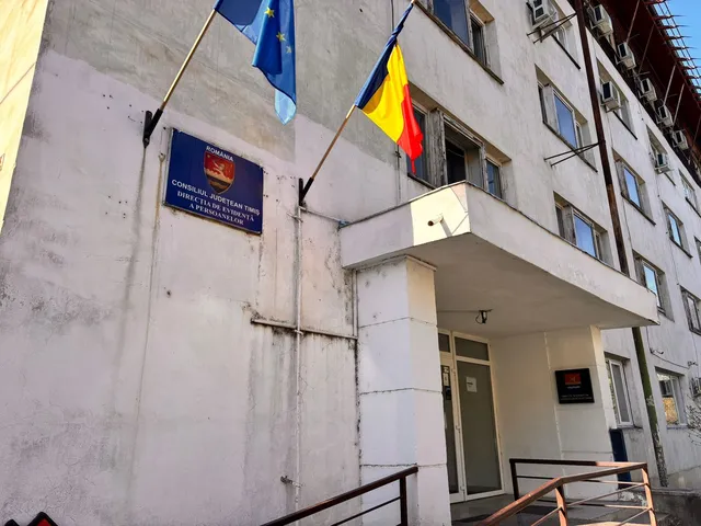 O clădire albă cu ferestre, un panou albastru pe perete și două steaguri, unul galben, roșu și albastru, celălalt albastru cu stele galbene.