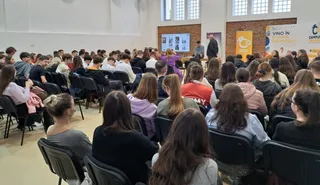 O mulțime de persoane stă pe scaune negre într-o sală, privind spre un ecran și un orator aflat în față.
