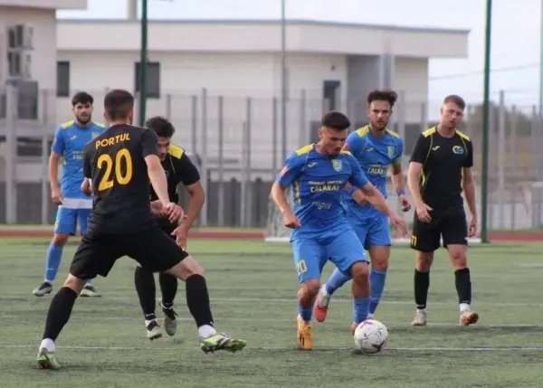 Cei doi jucători în echipament albastru concurează pentru un balon pe un teren de fotbal, în prezența altor jucători și a unui arbitru.