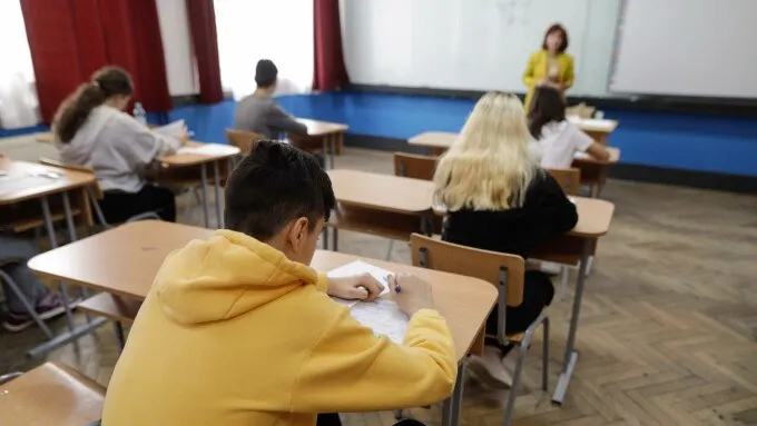 Elevi stau la bănci în interiorul unei săli de clasă, scriind pe foi de hârtie sub supravegherea unei persoane în picioare.