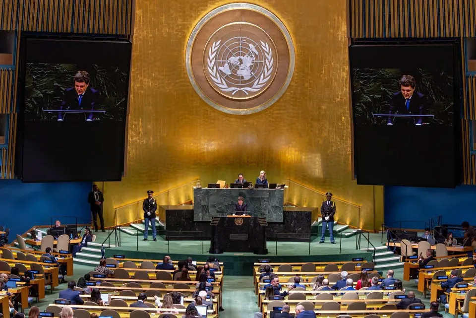 Un bărbat vorbește la un podium într-o sală de conferințe cu un simbol circular mare pe peretele din spate și ecrane laterale afișând imaginea sa.