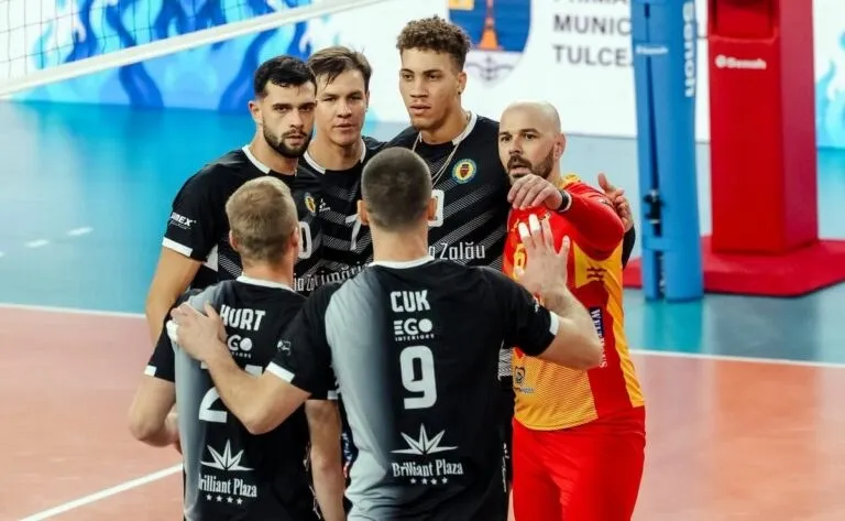 Cinci jucători de volei în echipamente negre și unul în echipament roșu și galben stau grupați pe un teren de sport.