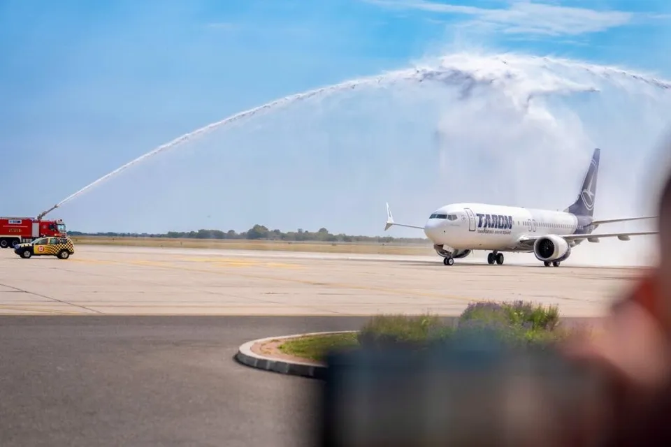 Un avion alb cu inscripția TAROM este stropit cu jeturi de apă de pe o pistă de decolare sub un cer senin.