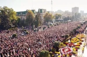 O mulțime mare de oameni este adunată pe o stradă largă lângă clădiri și copaci, lângă straturi de flori colorate.