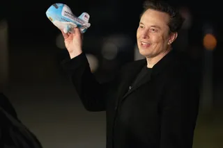 Elon Musk zâmbește, ținând ridicată în dreapta o jucărie de avion cu inscripții albastre și roșii.