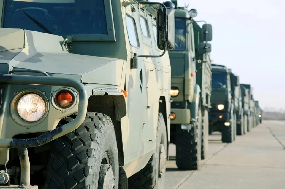 O coloană de vehicule militare de culoare verde este parcată pe un drum sub un cer luminos.