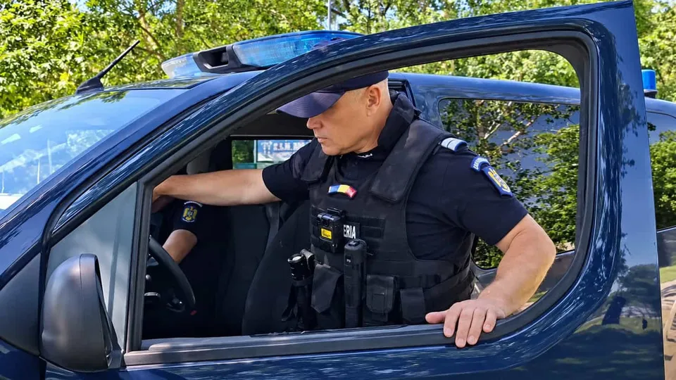 Un bărbat cu uniformă neagră și vestă tactică stă la volanul unei mașini albastre cu ușa deschisă.
