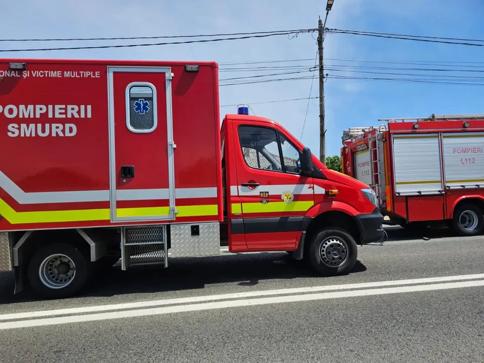 Un vehicul de serviciu roșu cu inscripția POMPIERII SMURD este parcat pe marginea unui drum asfaltat sub un cer senin.