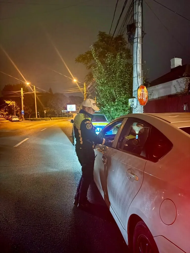 Un agent de poliție în uniformă stă lângă portiera deschisă a unei mașini albe pe un drum iluminat noaptea.