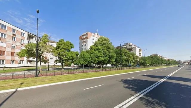 O stradă asfaltată cu linii albe traversează un spațiu verde cu copaci și clădiri de apartamente sub un cer senin.