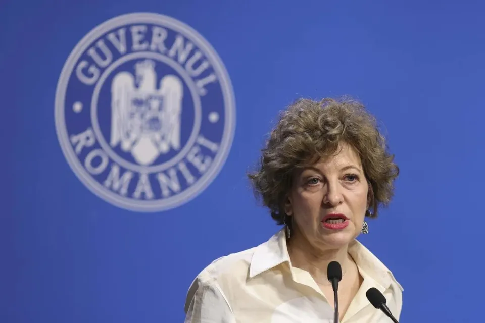 O femeie cu părul creț și cămașă deschisă la guler vorbește în fața unui microfon pe un fundal albastru cu un sigiliu circular.