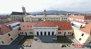O vedere aeriană a unei clădiri galbene cu acoperiș roșu și un turn de biserică înalt sub un cer înnorat.