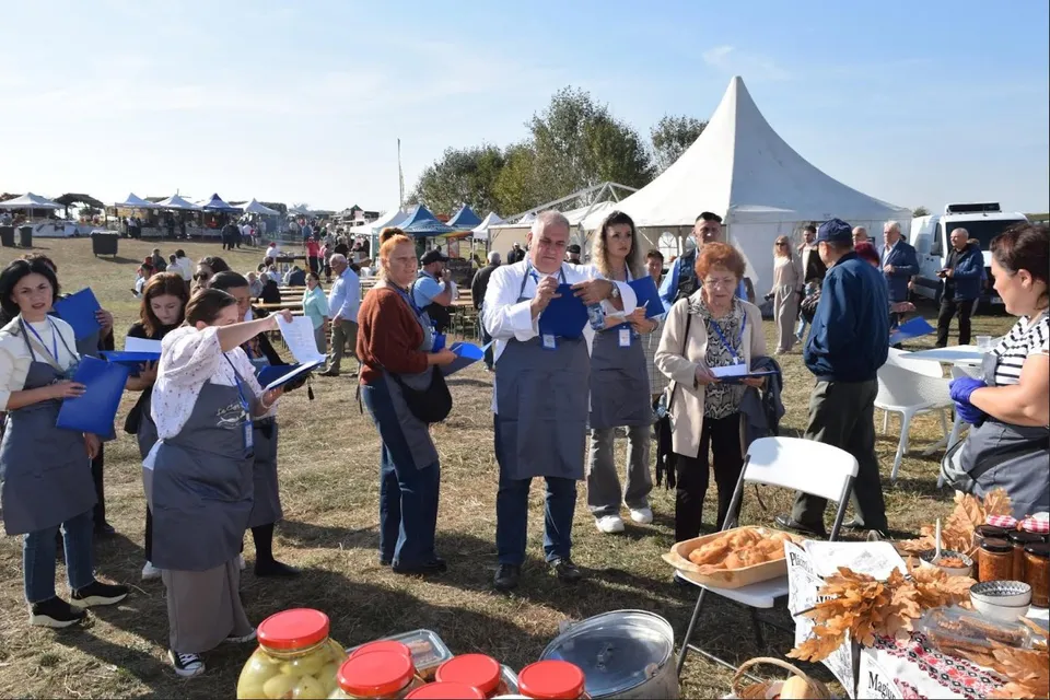 Un grup de oameni stă pe un câmp deschis lângă mese cu preparate culinare și corturi albe sub un cer senin.