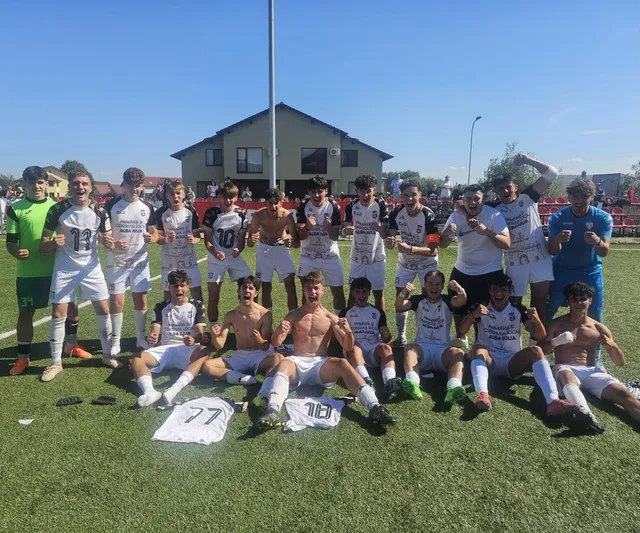 Un grup de tineri sportivi în echipamente de fotbal albe și colorate pozează pe un teren de fotbal verde sub un cer senin.
