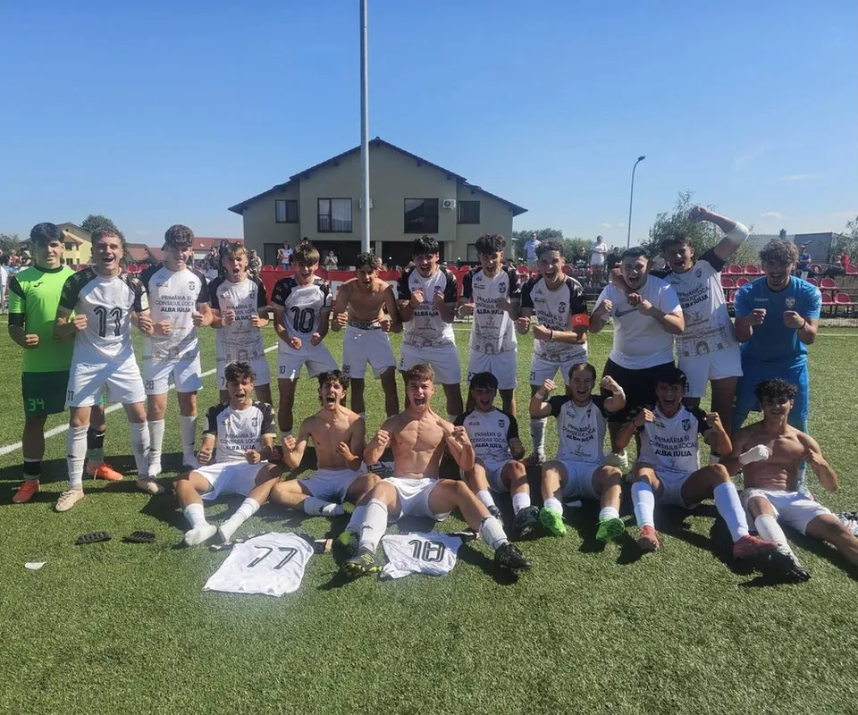 Un grup de tineri sportivi în echipamente de fotbal albe și colorate pozează pe un teren de fotbal verde sub un cer senin.