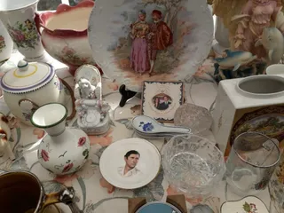 Diverse obiecte de ceramică și sticlă, inclusiv farfurii cu desene, vaze și căni, sunt dispuse pe o suprafață acoperită cu o față de masă decorată.