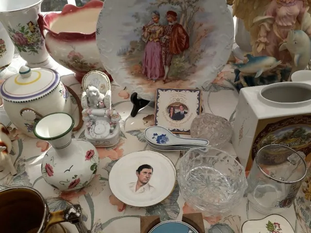 Diverse obiecte de ceramică și sticlă, inclusiv farfurii cu desene, vaze și căni, sunt dispuse pe o suprafață acoperită cu o față de masă decorată.
