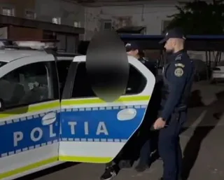 Un polițist în uniformă stă lângă o mașină de serviciu albastră și galbenă cu inscripția POLITIA, în timp ce un al doilea agent se află în spatele portierei deschise.