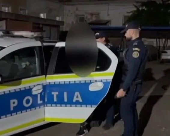Un polițist în uniformă stă lângă o mașină de serviciu albastră și galbenă cu inscripția POLITIA, în timp ce un al doilea agent se află în spatele portierei deschise.