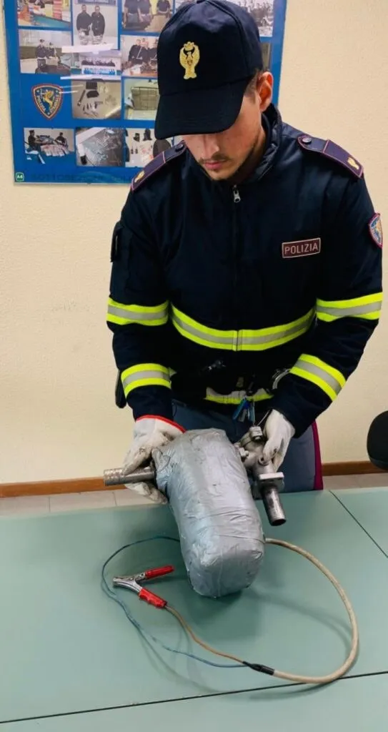 Un bărbat în uniformă de poliție cu benzi galbene se apleacă peste un obiect metalic acoperit cu folie argintie pe o masă.