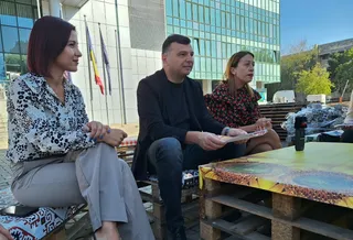 Trei persoane stau așezate pe paleți de lemn lângă o masă improvizată în fața unei clădiri moderne cu ferestre mari.