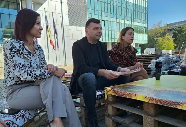 Trei persoane stau așezate pe paleți de lemn lângă o masă improvizată în fața unei clădiri moderne cu ferestre mari.