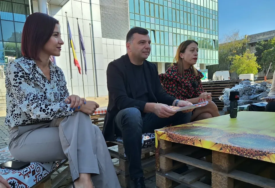 Trei persoane stau așezate pe paleți de lemn lângă o masă improvizată în fața unei clădiri moderne cu ferestre mari.
