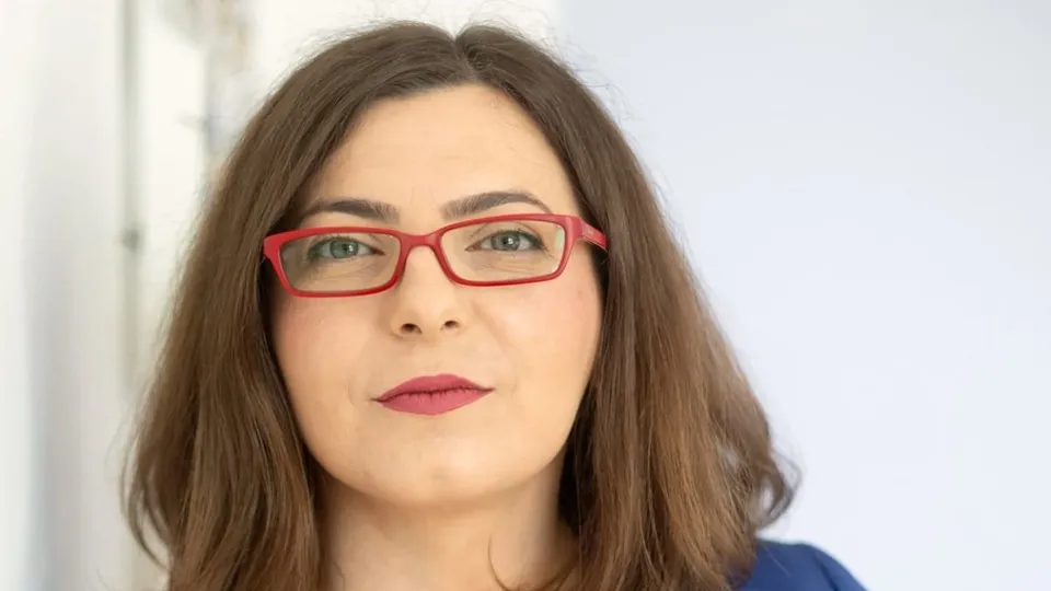 O femeie cu părul castaniu și ochelari cu rame roșii privește spre cameră.