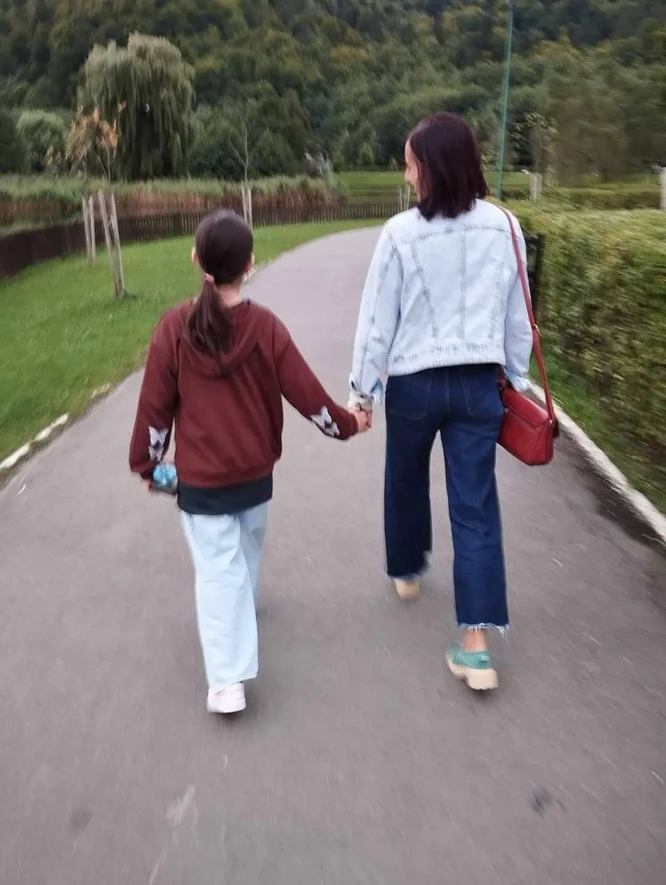Două persoane merg pe un drum asfaltat, o femeie cu bluză de denim și pantaloni închiși de mão în mâna unui copil cu hanorac bordo.