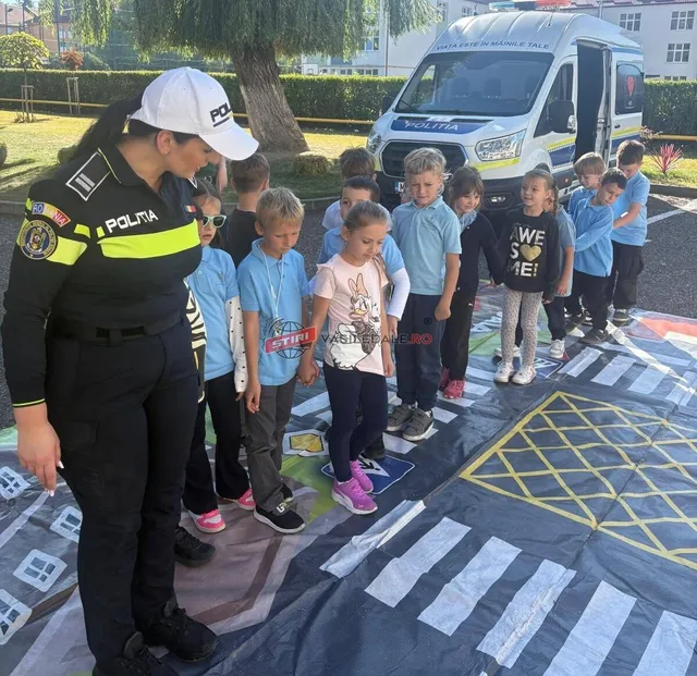 O polițistă în uniformă neagră și galbenă privește un grup de copii așezați pe un covor desenat cu marcaje rutiere colorate în exterior.