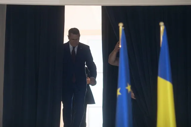 Un bărbat într-un costum închis și cravată merge printr-o deschidere între draperii negre, lângă două steaguri verticale.