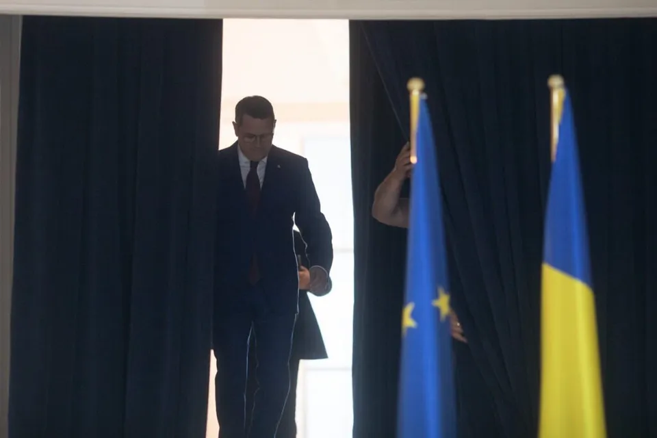 Un bărbat într-un costum închis și cravată merge printr-o deschidere între draperii negre, lângă două steaguri verticale.